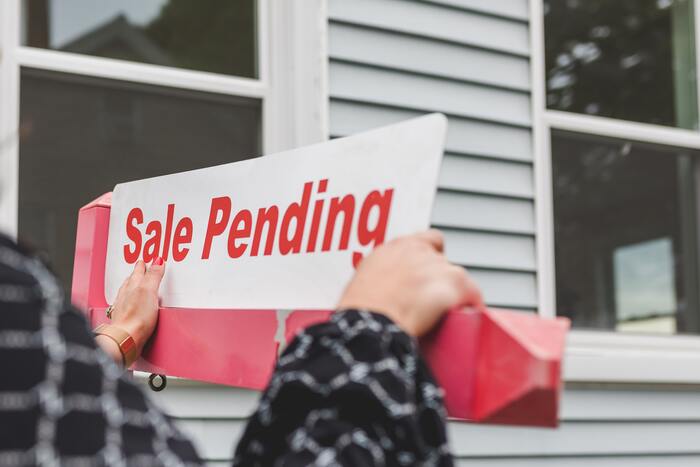The featured image for the article titled: "tax tips for property investors-selling your rental property". The image features a woman pasting a 'sale pending' sign outside of a home.